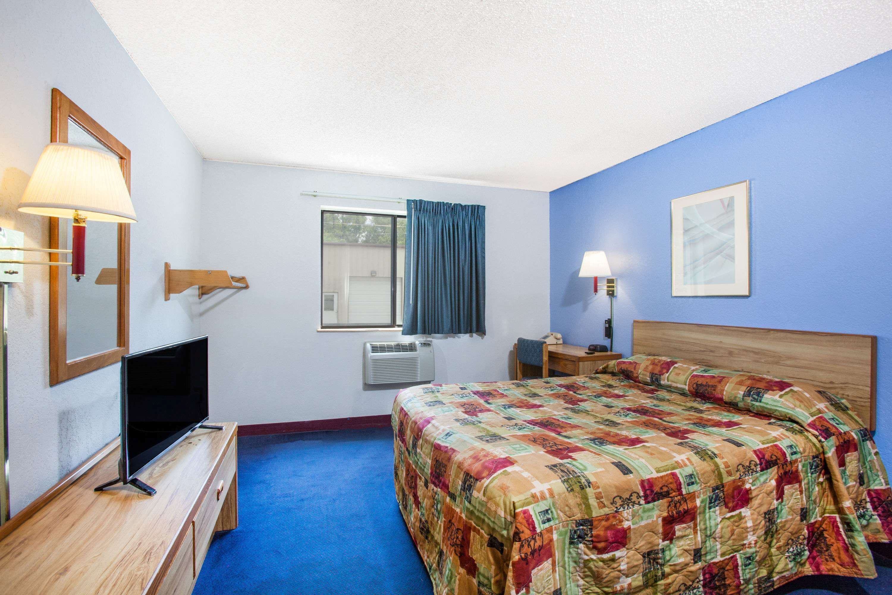 A hotel room with a wooden bed, a TV on a wooden dresser, a window with curtains, a wall-mounted lamp, and a framed picture.