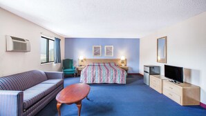 Habitación de hotel con sofá, cama con manta colorida, una mesita, televisor, escritorio con lámpara y espejo en la pared.