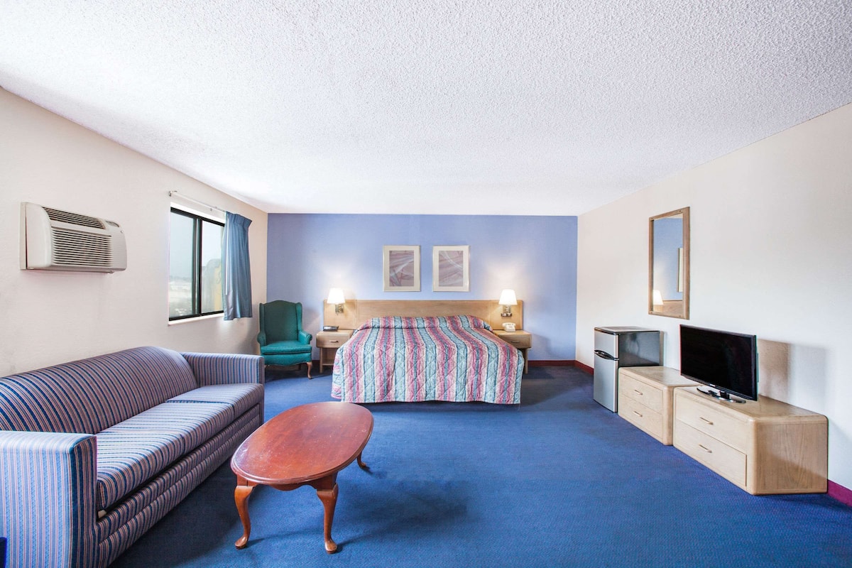 A hotel room with a sofa, a bed with a colorful blanket, a small table, a TV, a desk with a lamp, and a mirror on the wall.