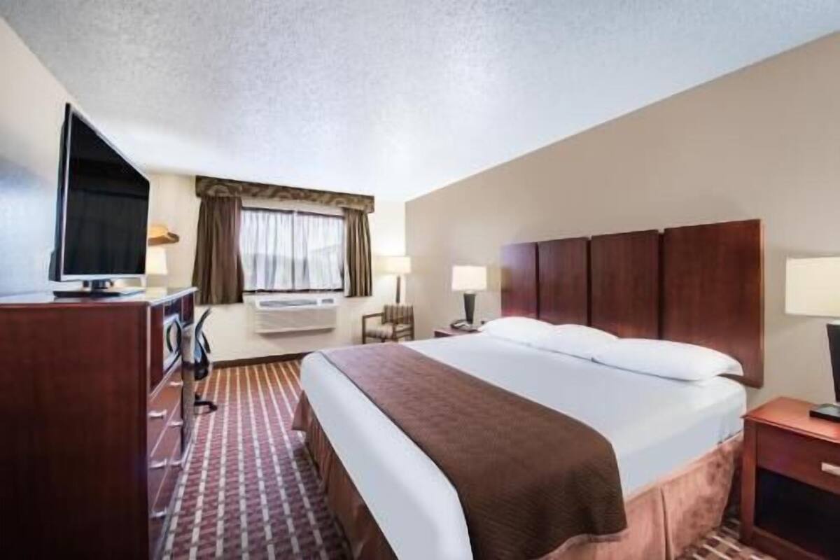 A hotel room with a bed, a desk with a television, a window with curtains, and a wall-mounted air conditioner.