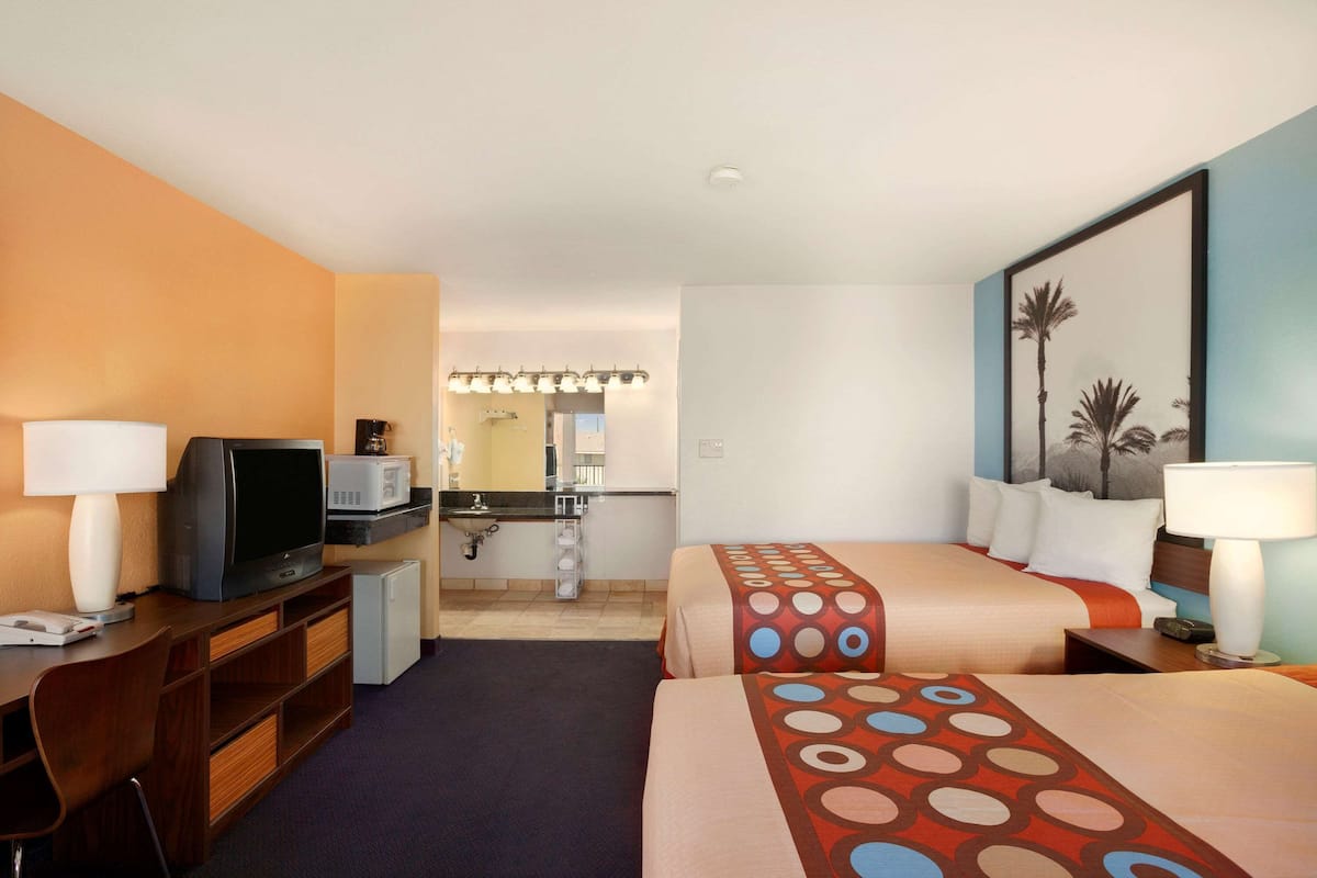 A hotel room with two beds, a desk with a television, a microwave, and a small kitchenette.