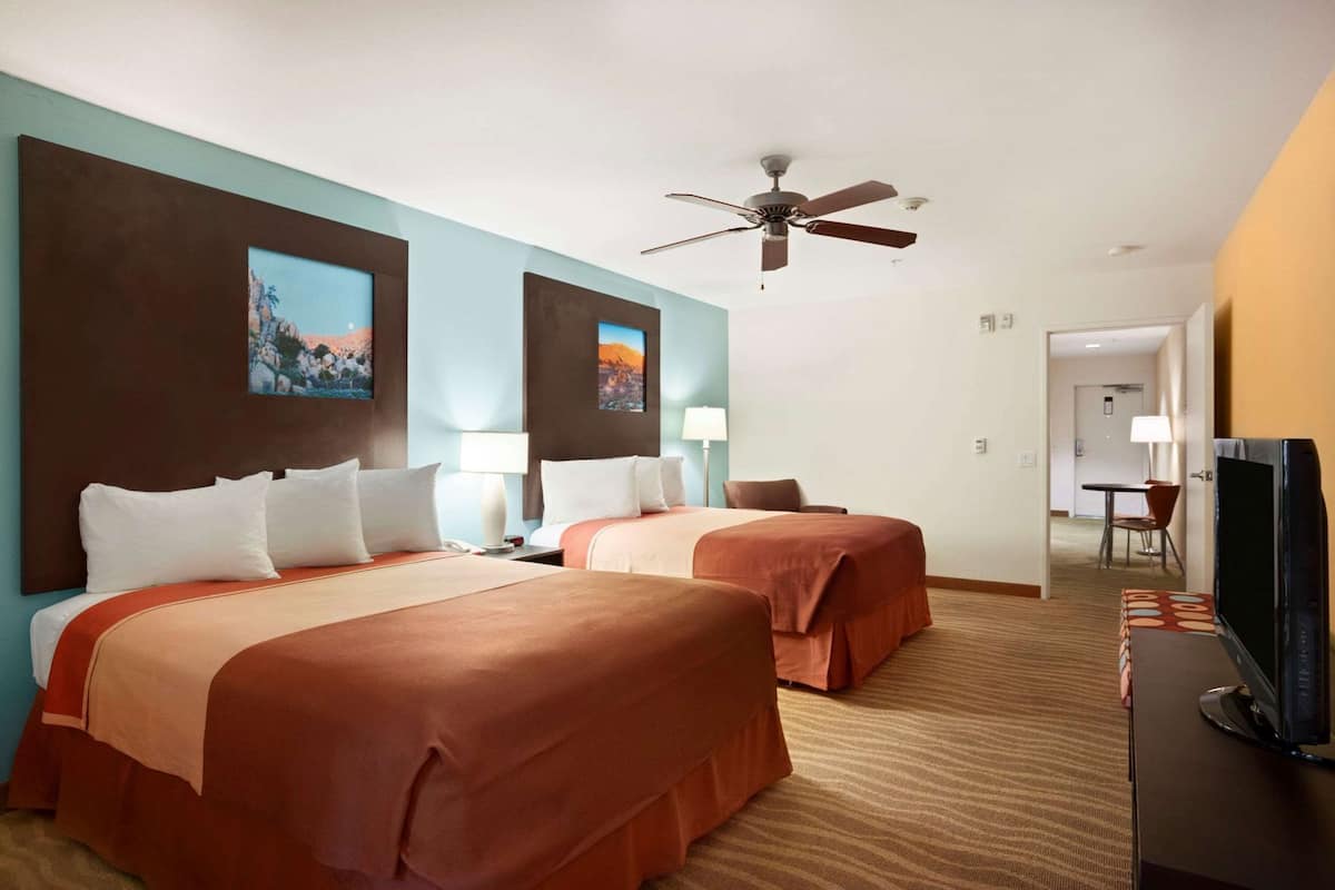 A hotel room with two beds, a ceiling fan, artwork on the walls, and a TV on a stand.
