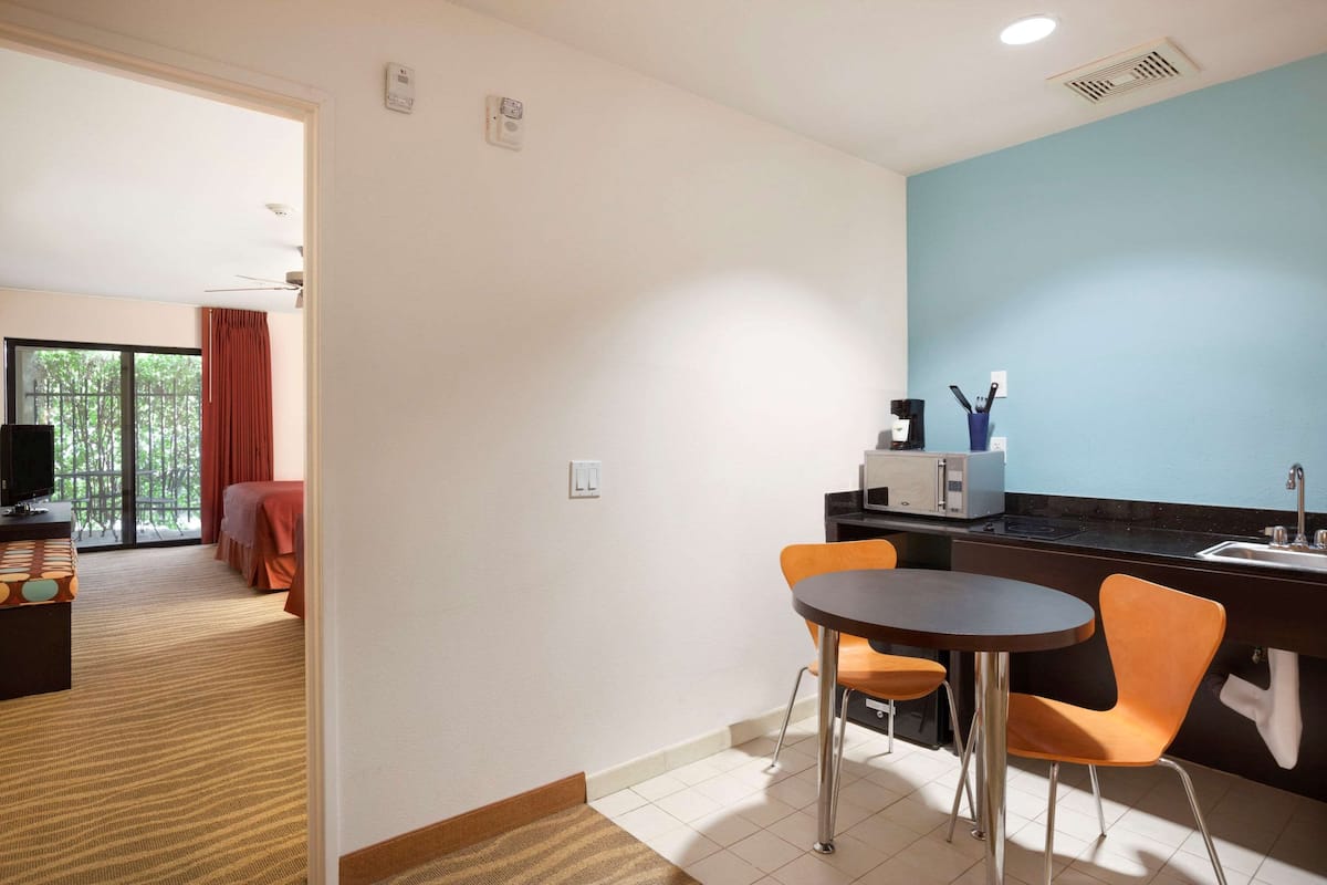A hotel room with a dining area featuring a round table and orange chairs, a kitchenette, and a view of a balcony with red curtains.