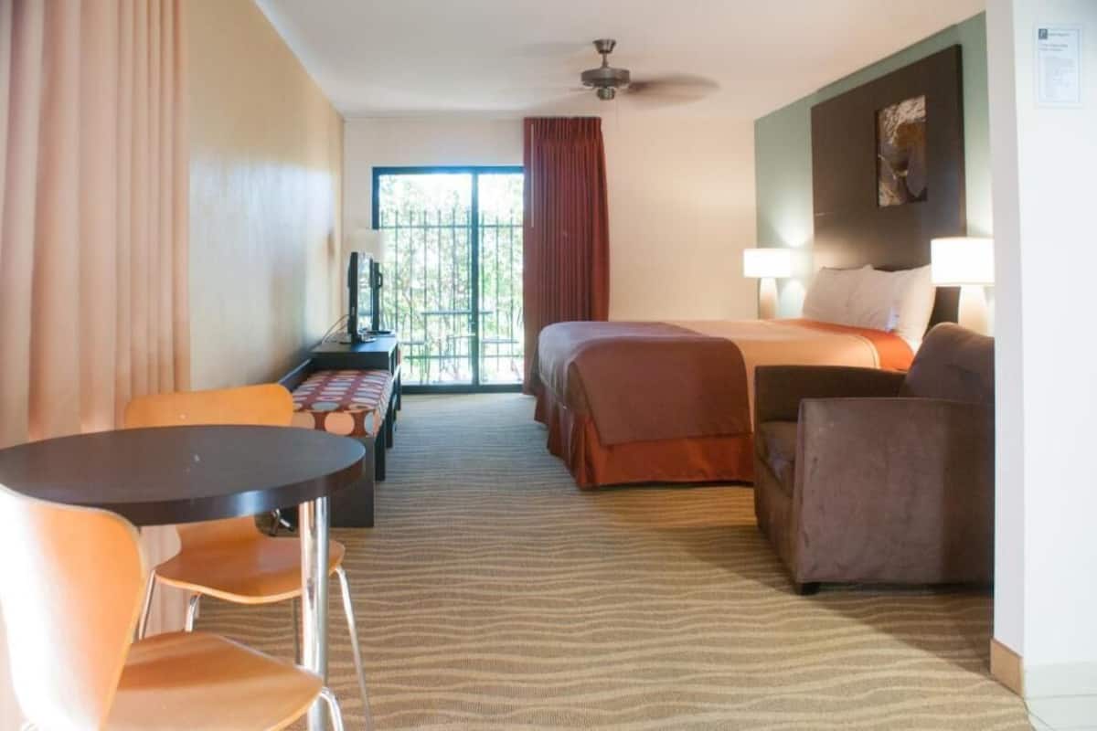 A hotel room with a bed, two bedside tables, a desk with a chair, a sofa, and a large window with curtains.