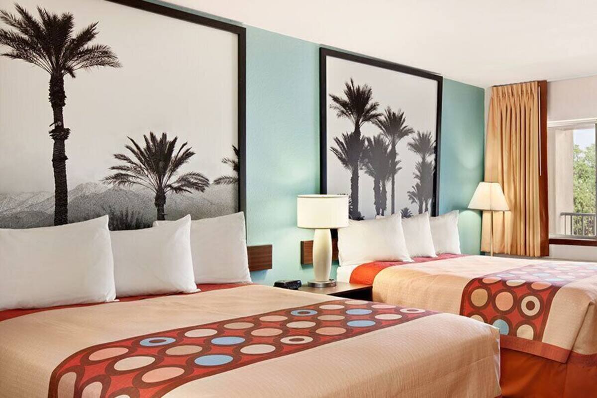 A hotel room with two beds, a chair, a window with curtains, and a painting of palm trees.