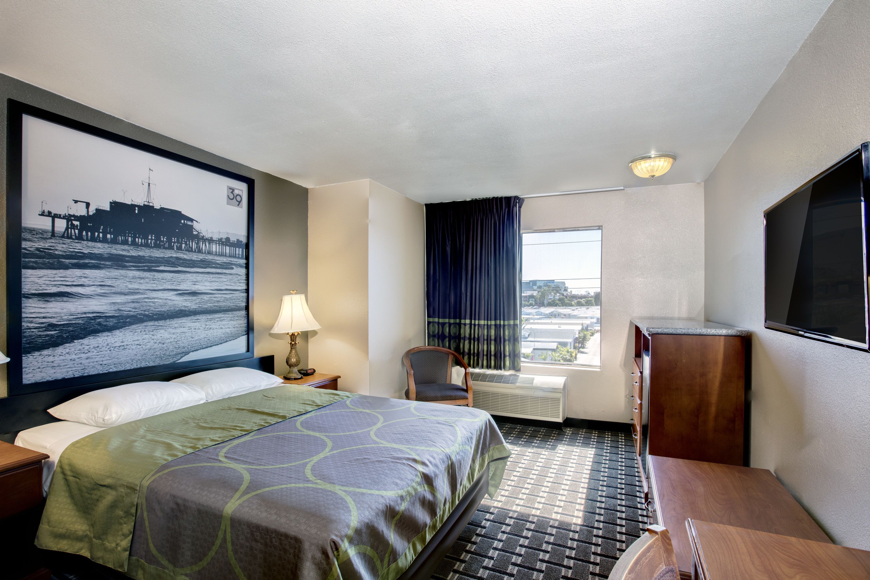 A hotel room with a bed, a desk, a chair, a TV, a window with curtains, and a large picture on the wall.