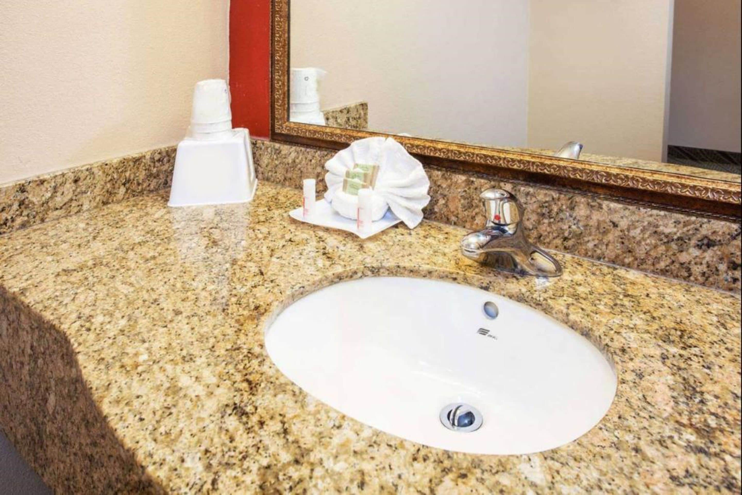 A granite countertop with a white sink and a faucet. There are toiletries on the counter, including a bottle and a small package. A mirror with a gold frame is above the sink.