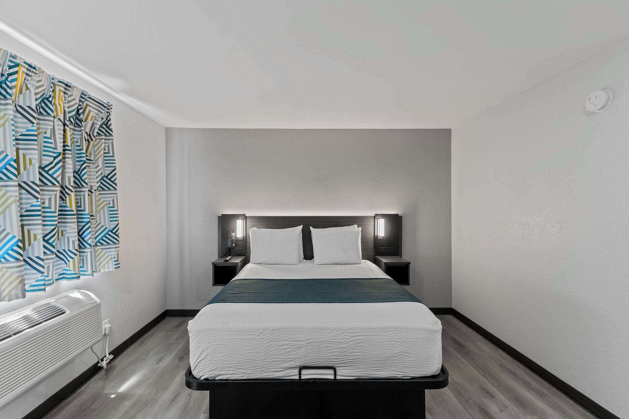 Una habitación de hotel con cama, mesitas de noche, un calentador en la pared y una cortina estampada.
