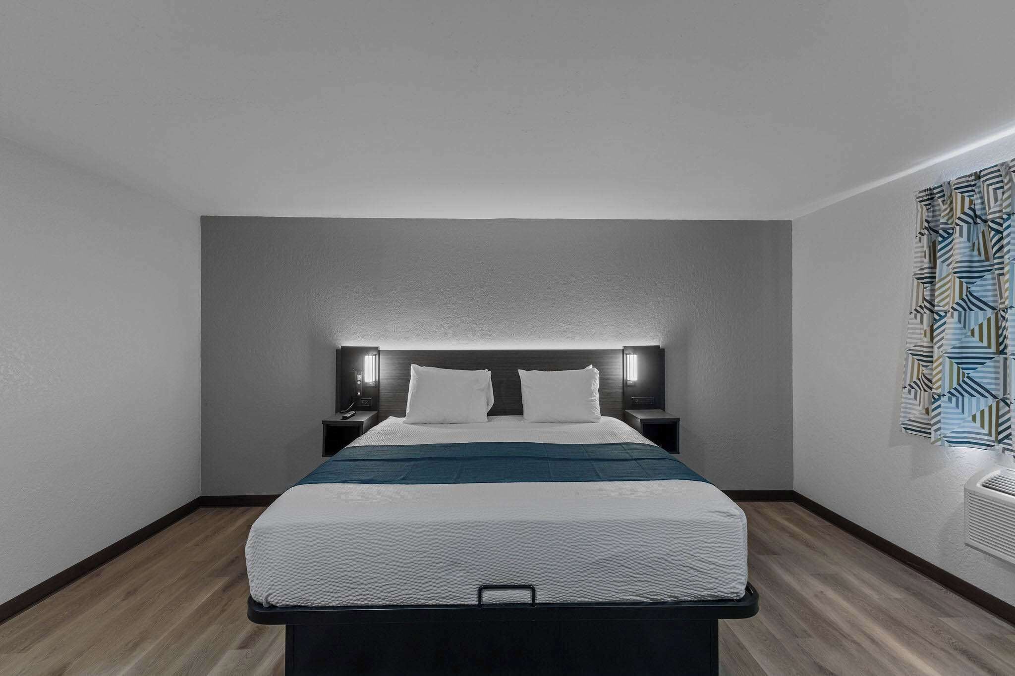 A modern hotel room with a large bed, a headboard with integrated lighting, a bedside table, and a patterned wall decoration.