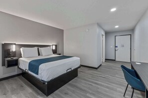 A modern hotel room with a bed, bedside table, and a desk with a chair. - Motel 6 Brighton CO Denver Northeast (Brighton)
