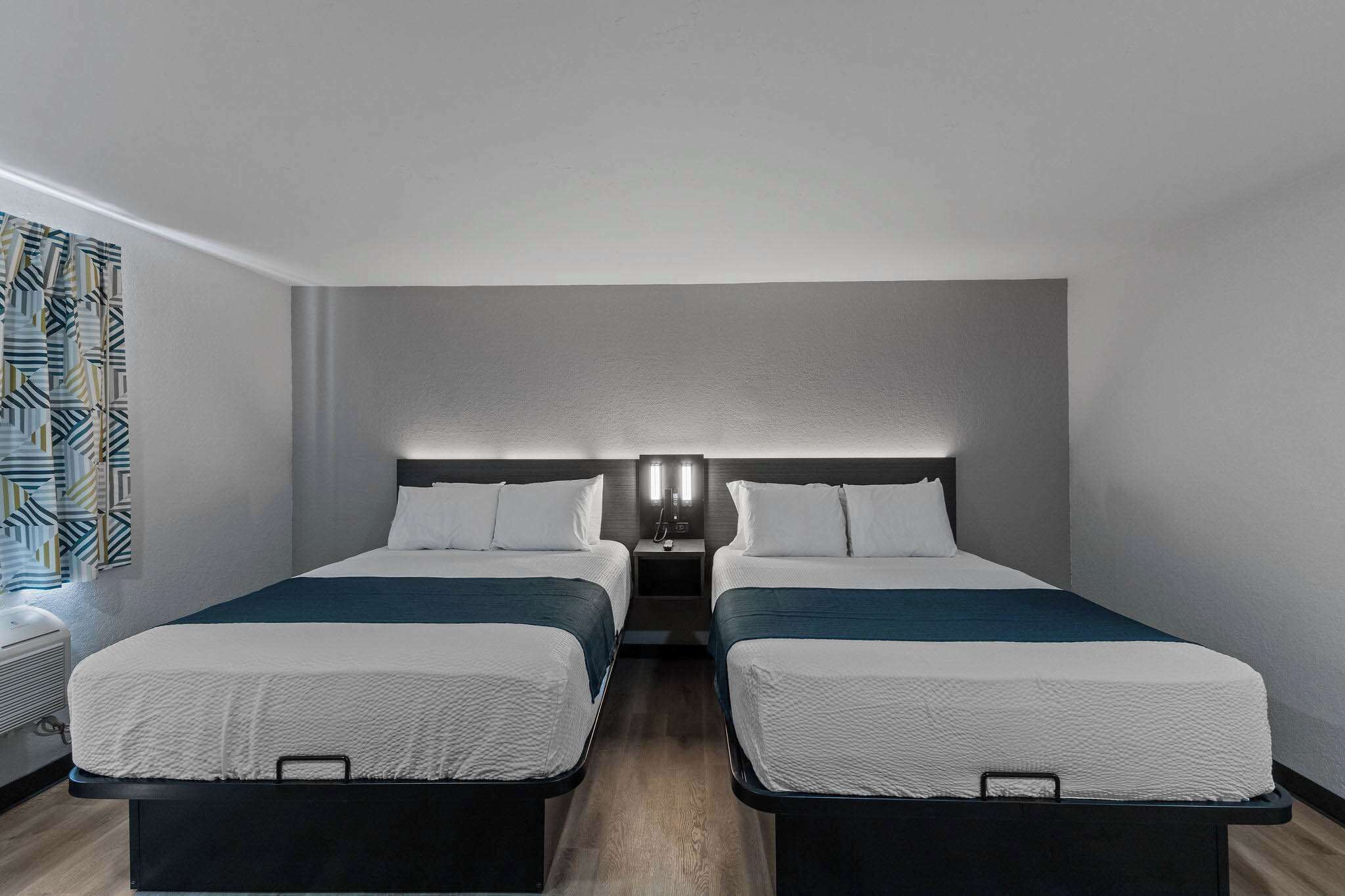 A hotel room with two single beds, a nightstand with a lamp, and a patterned curtain.
