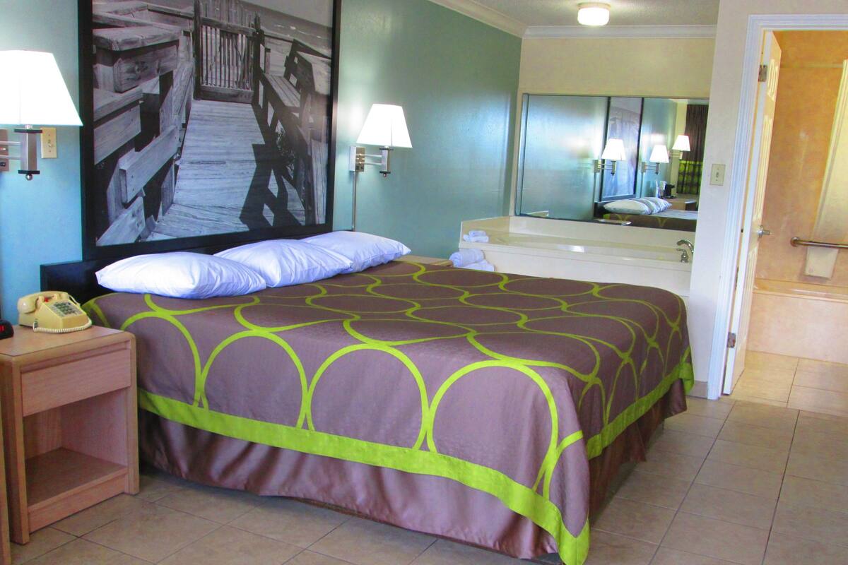 Une chambre d’hôtel avec un lit, une table de chevet avec un téléphone, un miroir et une salle de bain visible à travers une porte ouverte.