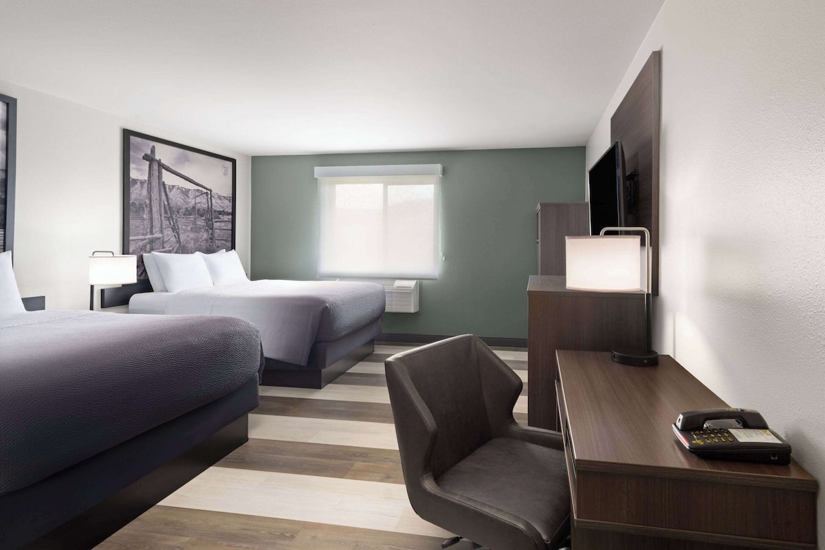 Habitación de hotel con dos camas, un escritorio con teléfono, una silla, un televisor y una ventana con persianas.