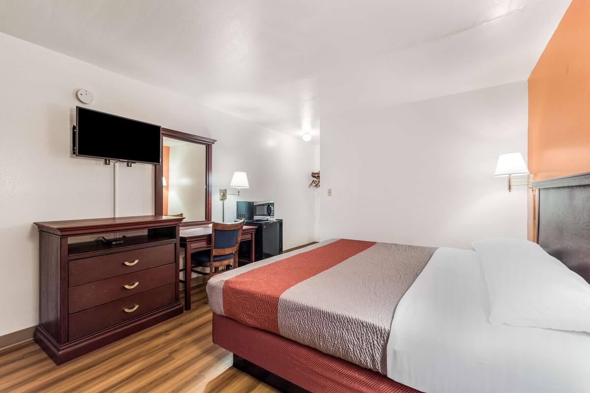 A hotel room with a bed, a desk, a chair, a dresser, a television, and a lamp.