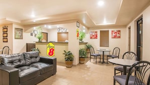 Un vestíbulo de hotel con un sofá de cuero negro, un mostrador de recepción, una zona de comedor con una mesa redonda y sillas, y plantas en macetas.
