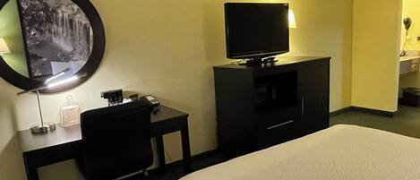 Habitación de hotel con una cama, un escritorio con silla, un televisor y un espejo montado en la pared.