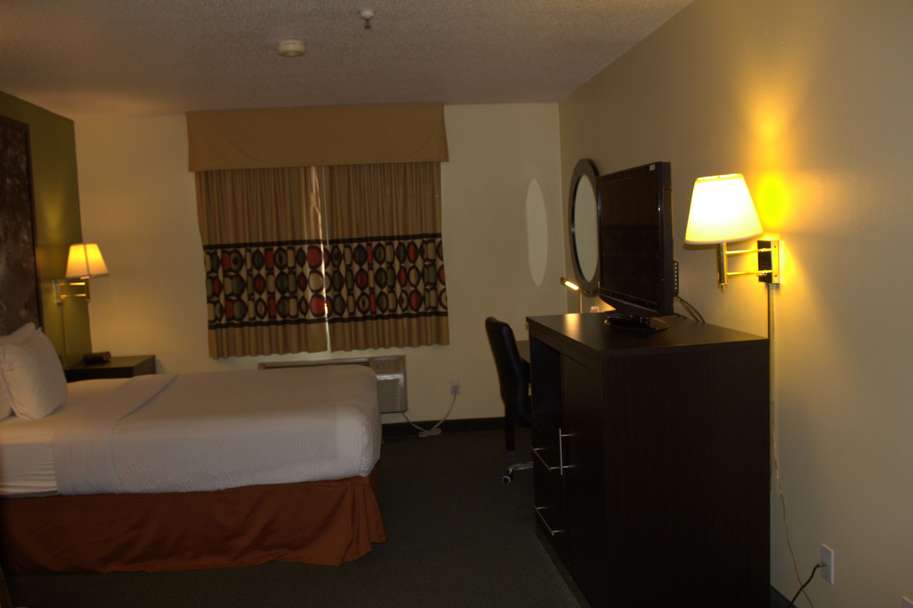Quarto de hotel com cama, escrivaninha, cadeira, televisão e duas luminárias de parede.