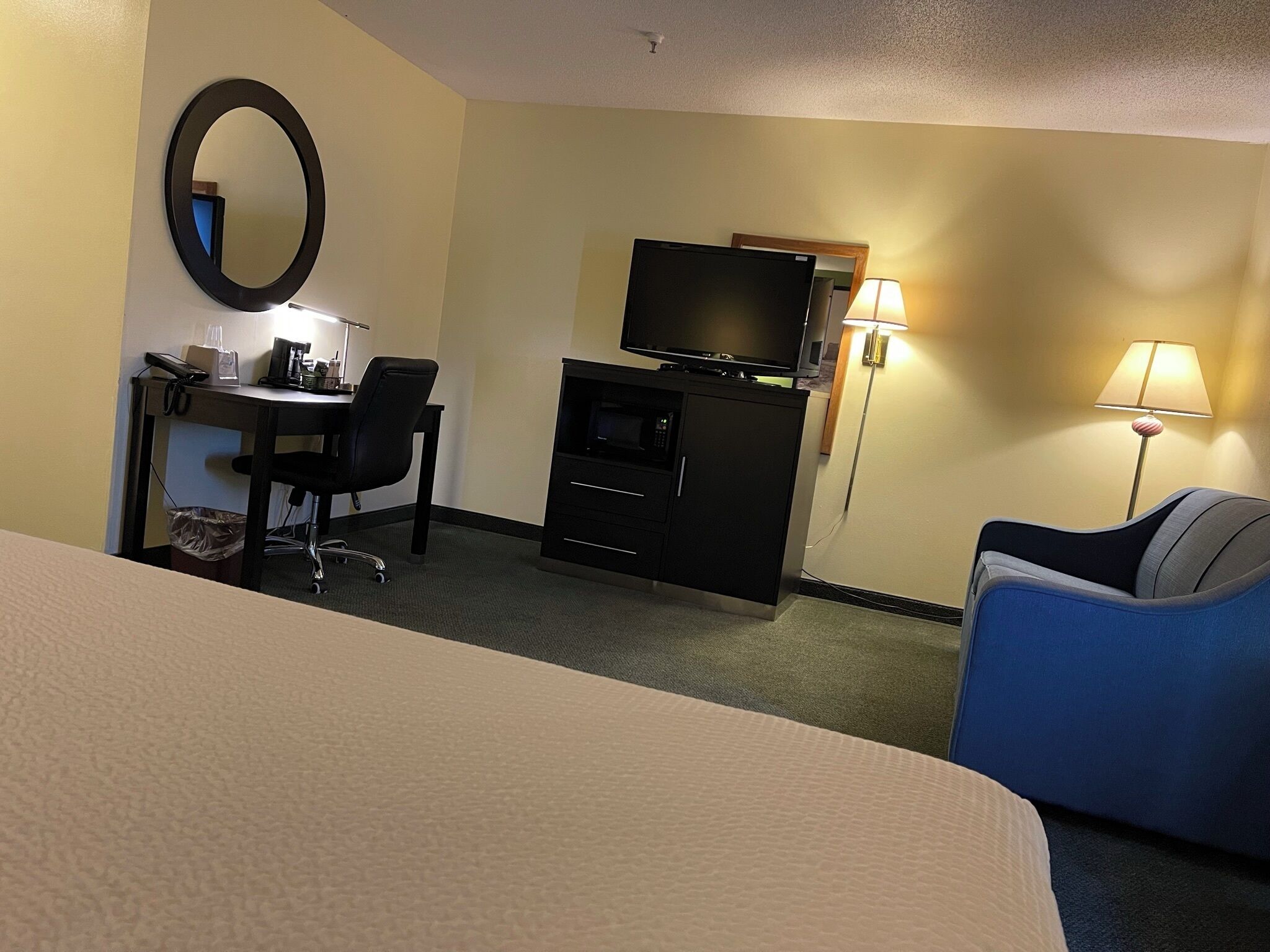 Habitación de hotel con cama, escritorio con silla, televisión, kitchenette pequeña y dos lámparas.