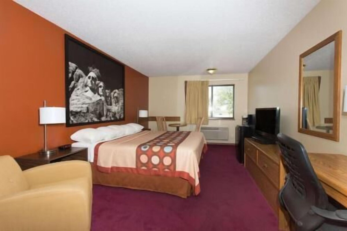 A hotel room with a bed, a desk, a chair, a TV, a window with curtains, and a painting on the wall.