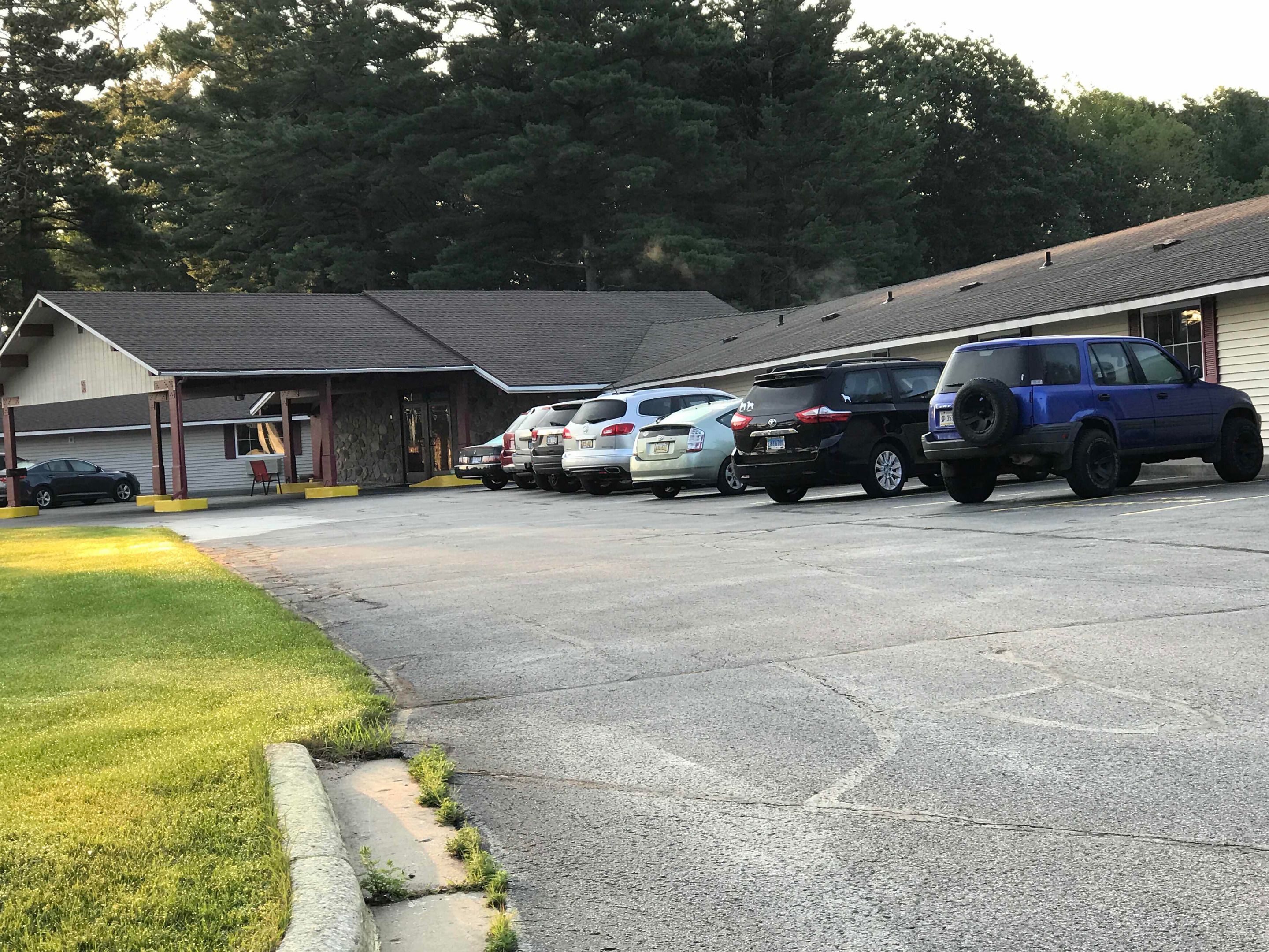 Un parking avec plusieurs voitures, un bâtiment doté d’une zone abritée et une pelouse avec un trottoir.