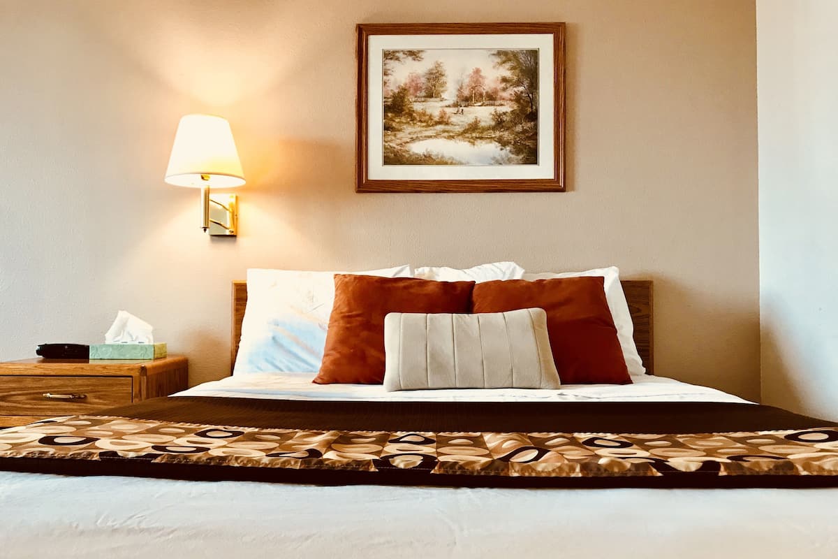 A neatly made bed with a patterned bedspread, a bedside lamp, a framed picture on the wall, and a wooden nightstand with a tissue box.