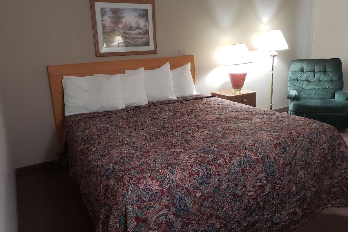 A hotel room with a large bed, a green armchair, a nightstand with a lamp, and a framed picture on the wall.