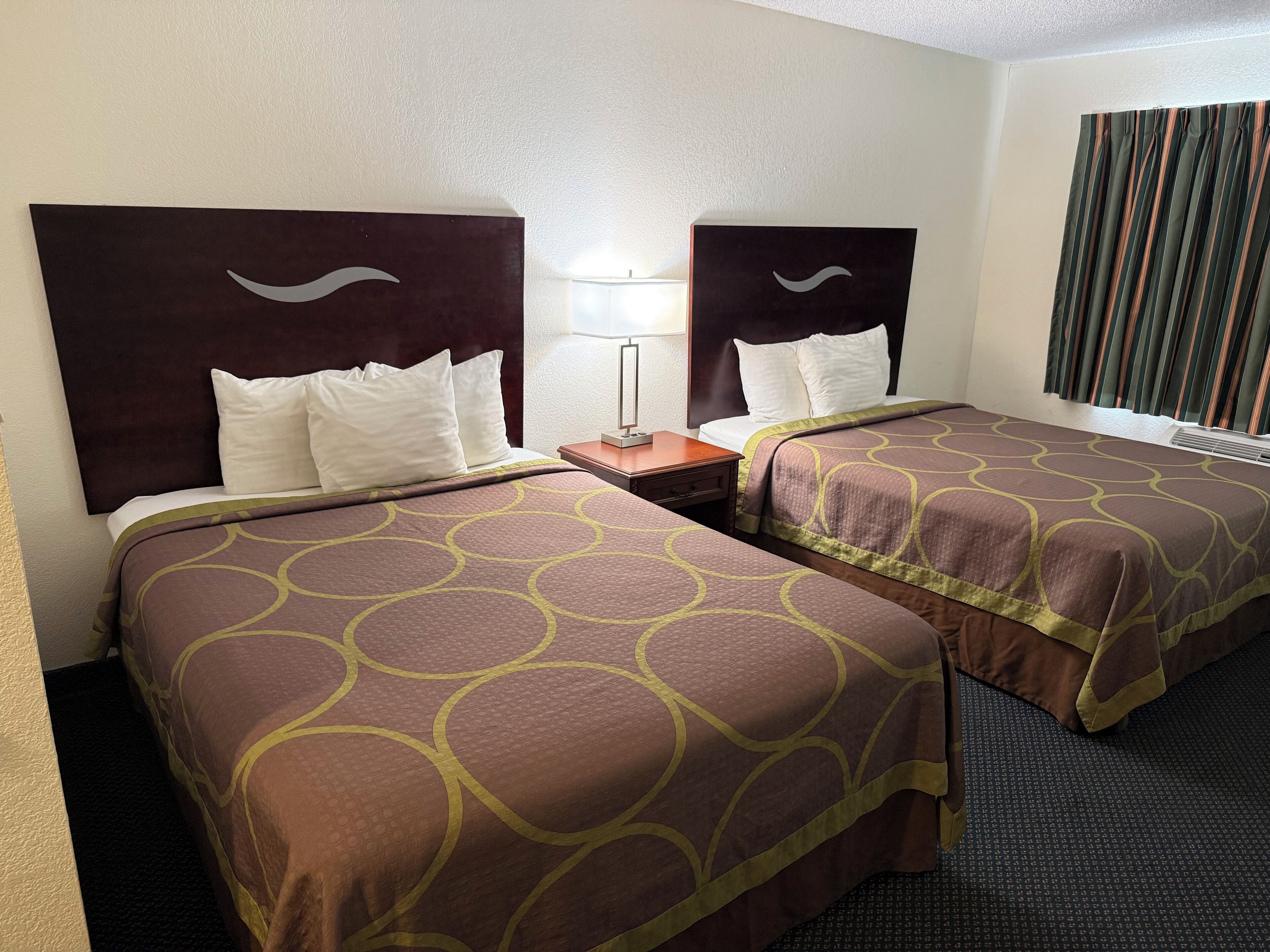 A hotel room with two beds, each with a headboard featuring a wave design, white pillows, and brown bedding with a circular pattern.