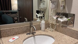 Une salle de bain d’hôtel avec un lavabo doté d’un plan de travail en granit, un miroir, un porte-serviettes avec des serviettes pliées, un sèche-cheveux et un distributeur de gel hydroalcoolique.