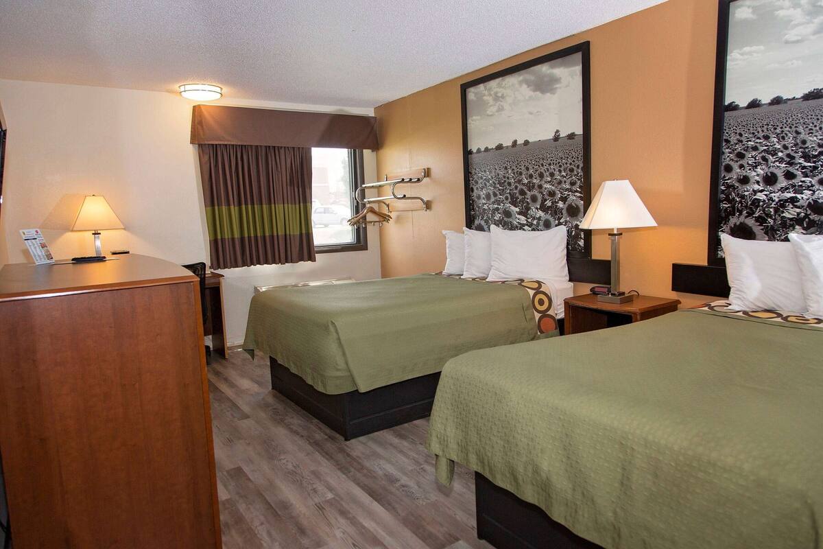 A hotel room with two beds, a desk, a chair, a window with curtains, and a wall-mounted lamp.