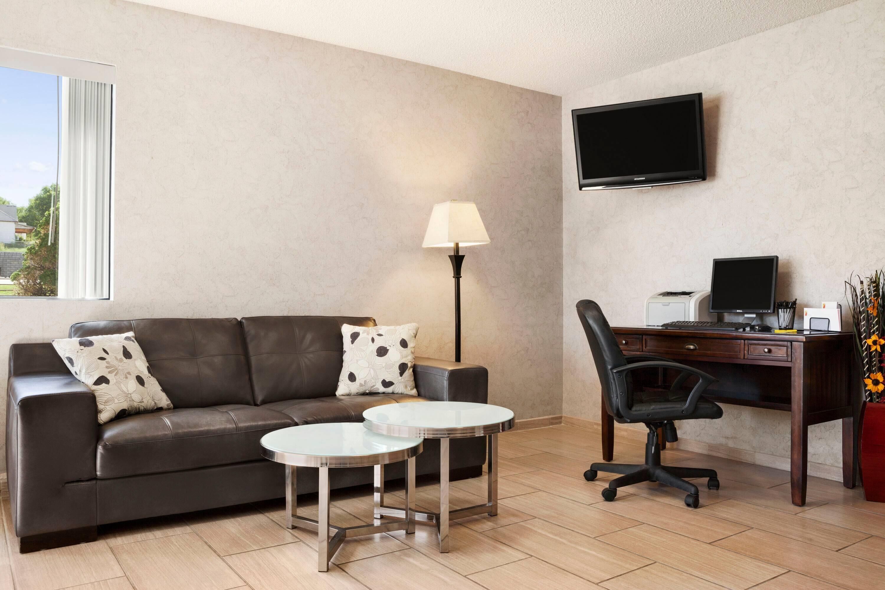A modern hotel room with a leather sofa, two round tables, a desk with a computer, a lamp, and a TV on the wall.