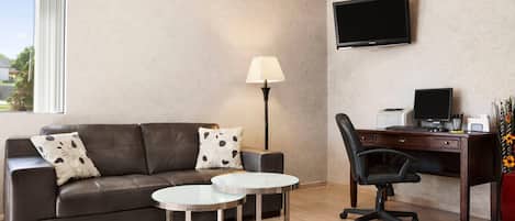 Una habitación de hotel moderna con un sofá de cuero, dos mesas redondas, un escritorio con computadora, una lámpara y un televisor en la pared.