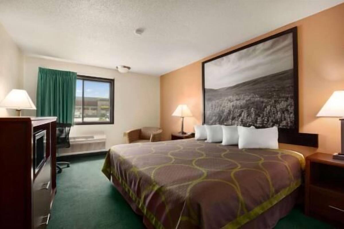 A hotel room with a bed, two bedside lamps, a desk, a chair, a window with curtains, and a large painting on the wall.