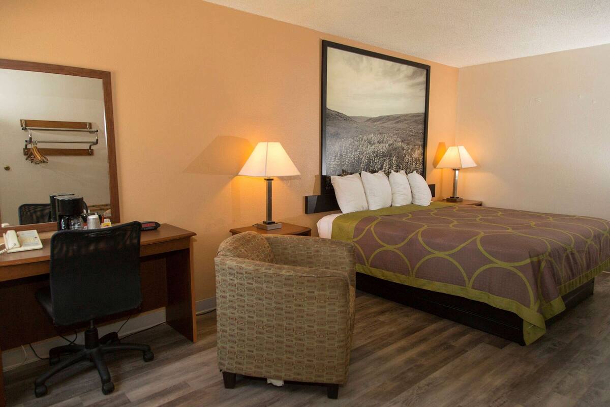 A hotel room with a bed, two bedside lamps, a desk with a chair, a small armchair, and a large framed picture on the wall.