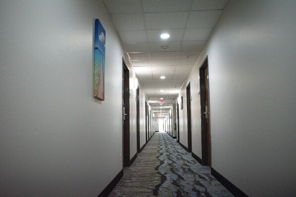 A long, narrow hotel corridor with doors on both sides, a patterned carpet, and a ceiling with recessed lighting. - Super 8 by Wyndham Pratt (Pratt)