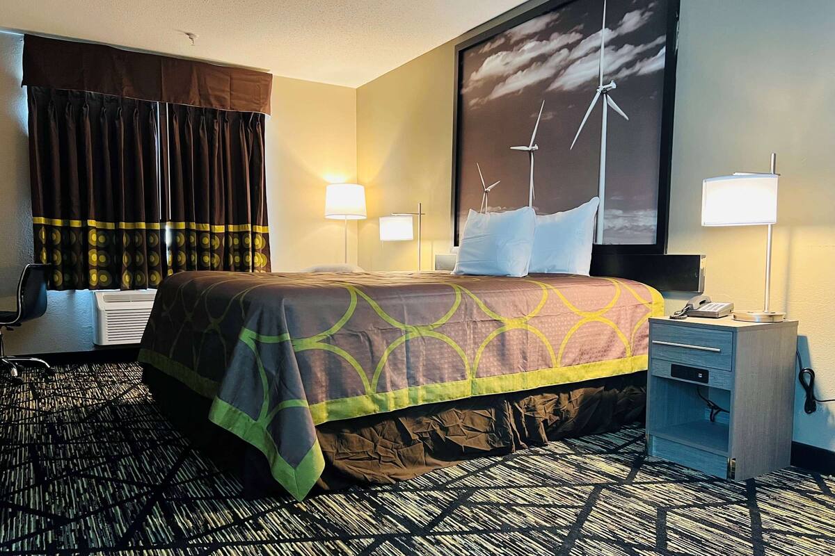 Une chambre d’hôtel comprenant un lit, un bureau, une chaise, une lampe et un mur orné d’un tableau représentant une éolienne.