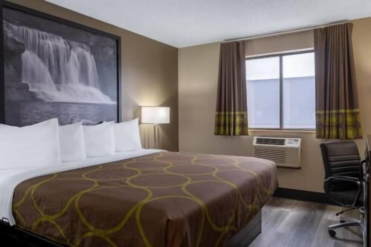 Habitación de hotel con cama, una silla, ventana con cortinas, unidad de aire acondicionado y un cuadro colgado en la pared.