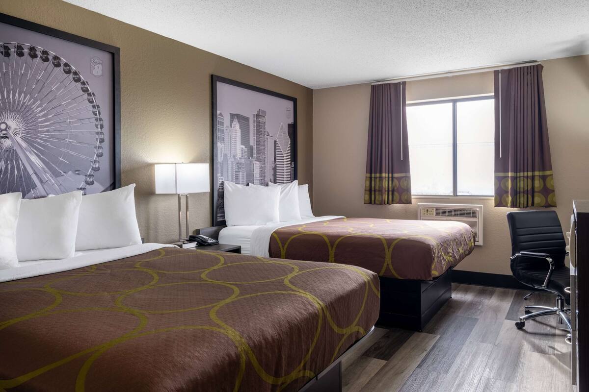 Habitación de hotel con dos camas, un escritorio, una silla, un ventanal con cortinas y una fotografía de un paisaje urbano en la pared.