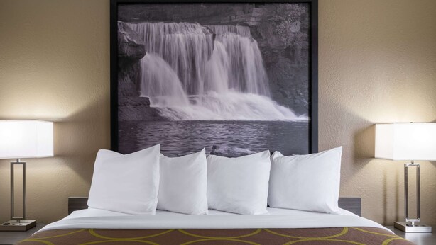 Une chambre d’hôtel avec un lit, des oreillers blancs, un tableau représentant une cascade, et deux lampes de chevet.