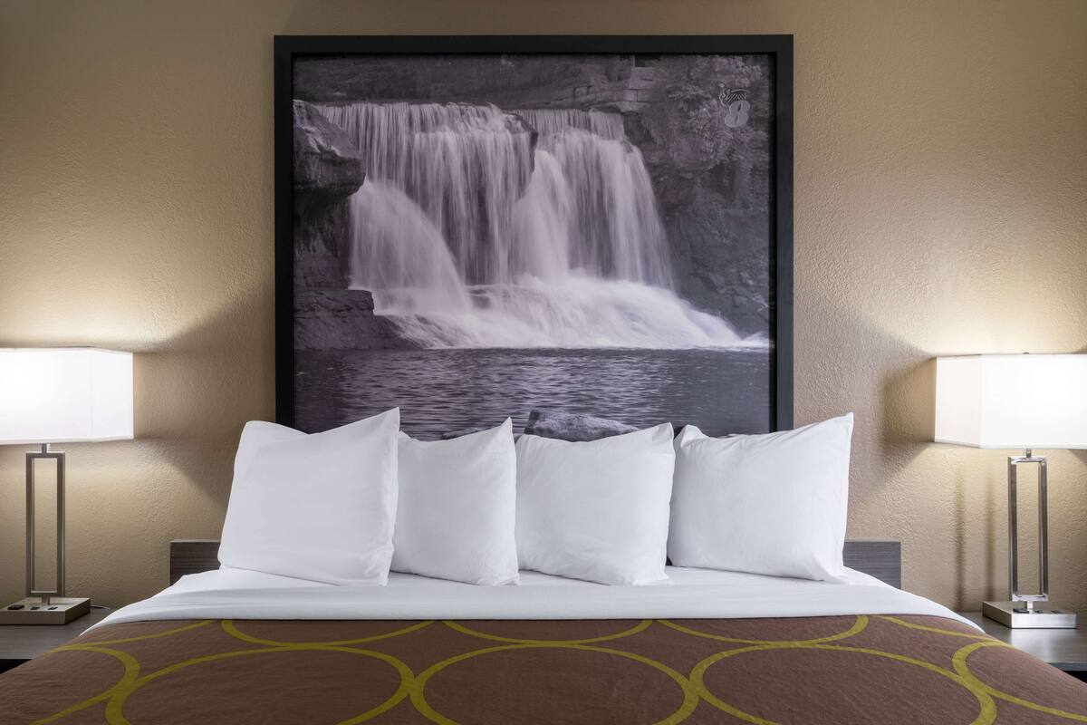 Una habitación de hotel con una cama, almohadas blancas, un cuadro enmarco de una cascada y dos lámparas de noche.