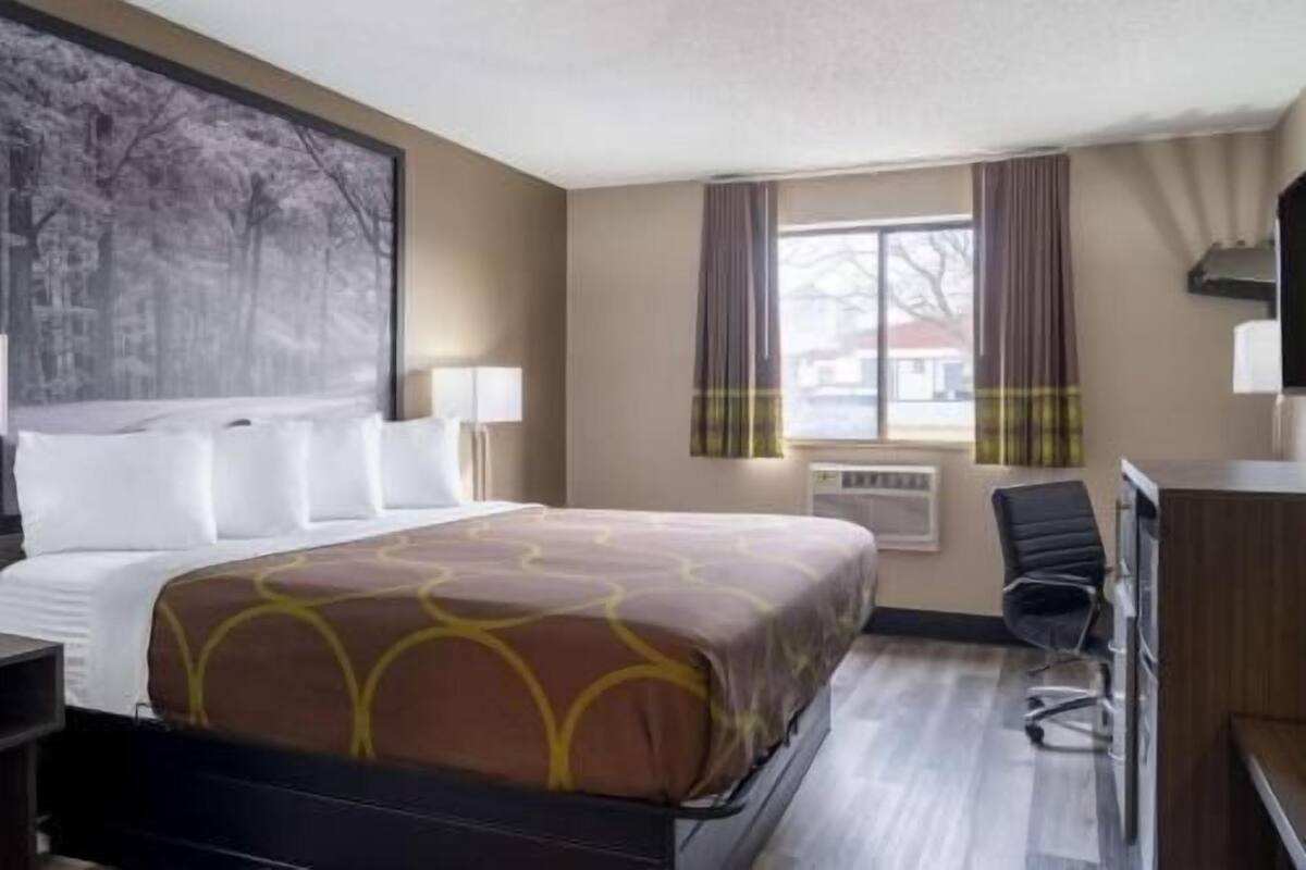 Un cuarto de hotel con una cama grande, un escritorio, una silla, una ventana con cortinas y un cuadro en la pared.