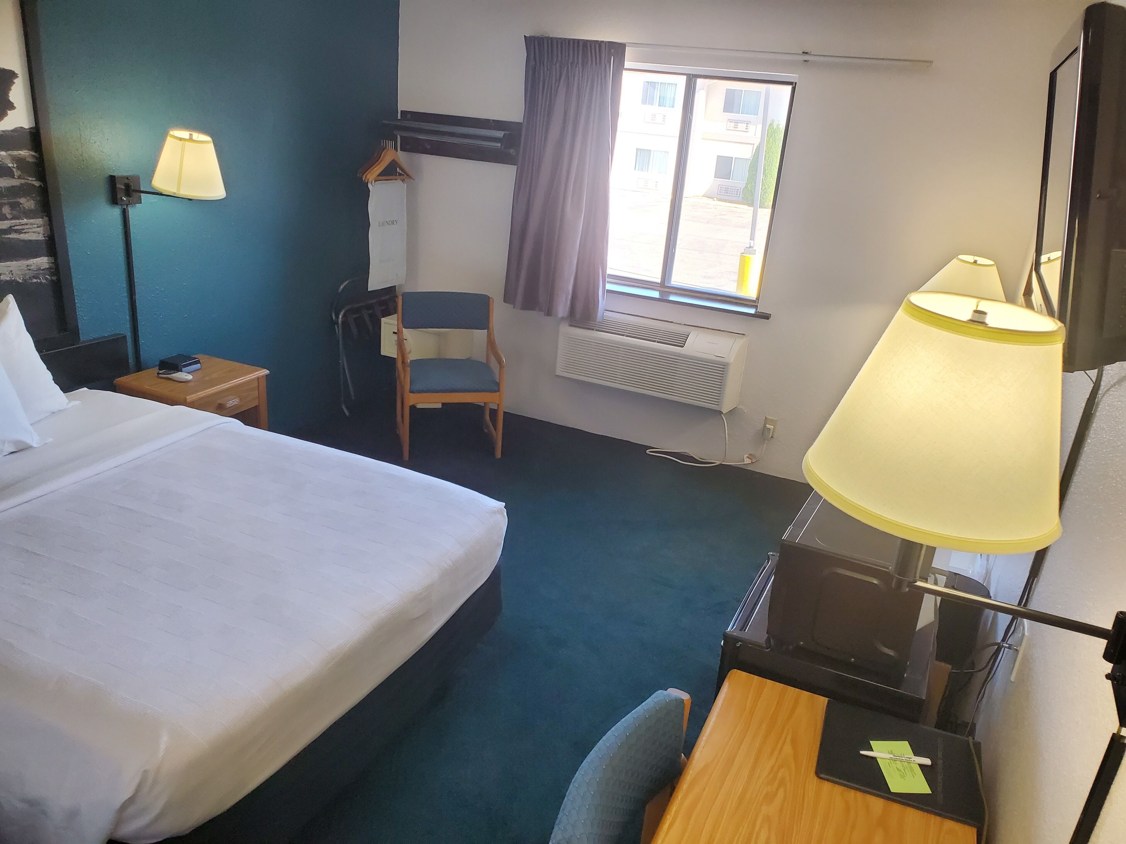 Ein Hotelzimmer mit einem Bett, einem Schreibtisch, einem Sessel, einer Schreibtischlampe, einem Fenster mit Vorhängen, einer Klimaanlage und einem Wandtuchhalter.