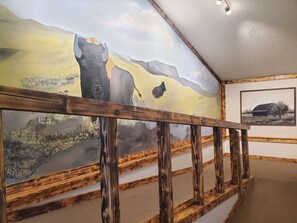 A room with a mural of a bull and a landscape, a wooden railing, and a framed picture on the wall. - Glendive Lodge (Glendive)
