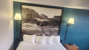 Une chambre d’hôtel avec un lit, deux lampes fixées au mur, une table de chevet avec une télécommande, et un grand tableau représentant un paysage de montagne au-dessus du lit.