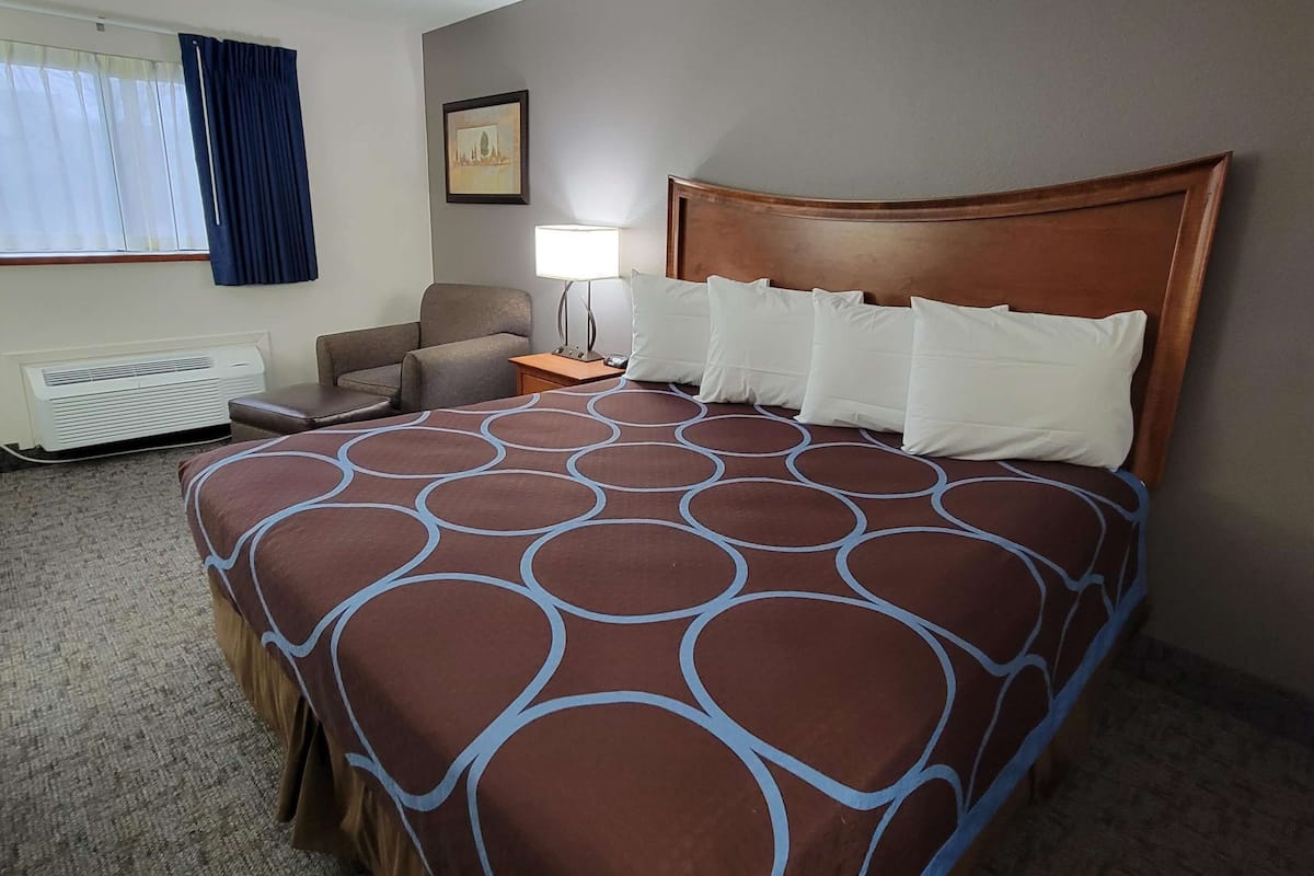 Een hotelkamer met een groot bed, een nachtkastje met lamp, een stoel, een radiator en een raam met gordijnen.