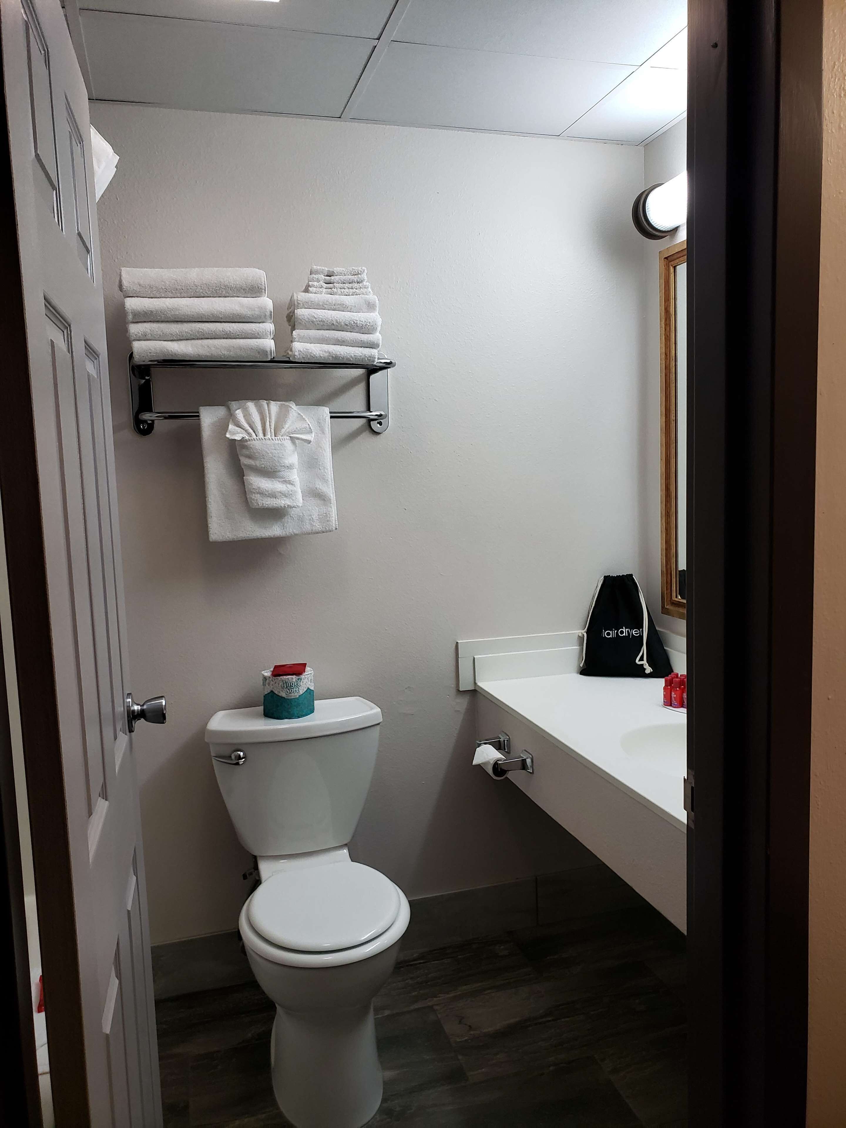 A bathroom with a toilet, a towel rack with folded towels, a toilet paper holder, a sink with a mirror, and a black bag labeled "hair dryer".