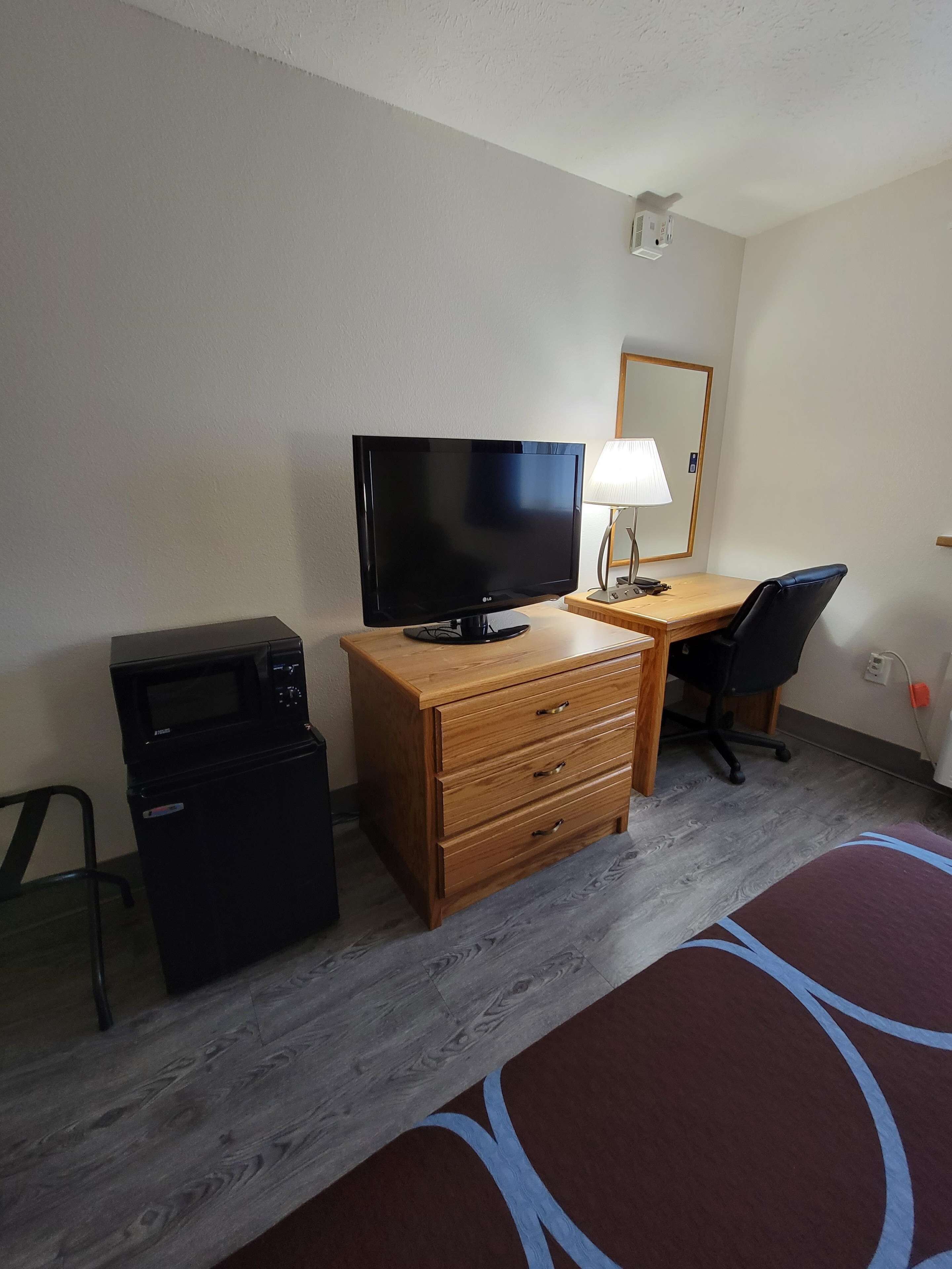 A hotel room with a wooden desk, a flat-screen TV, a lamp, a microwave, and a chair.