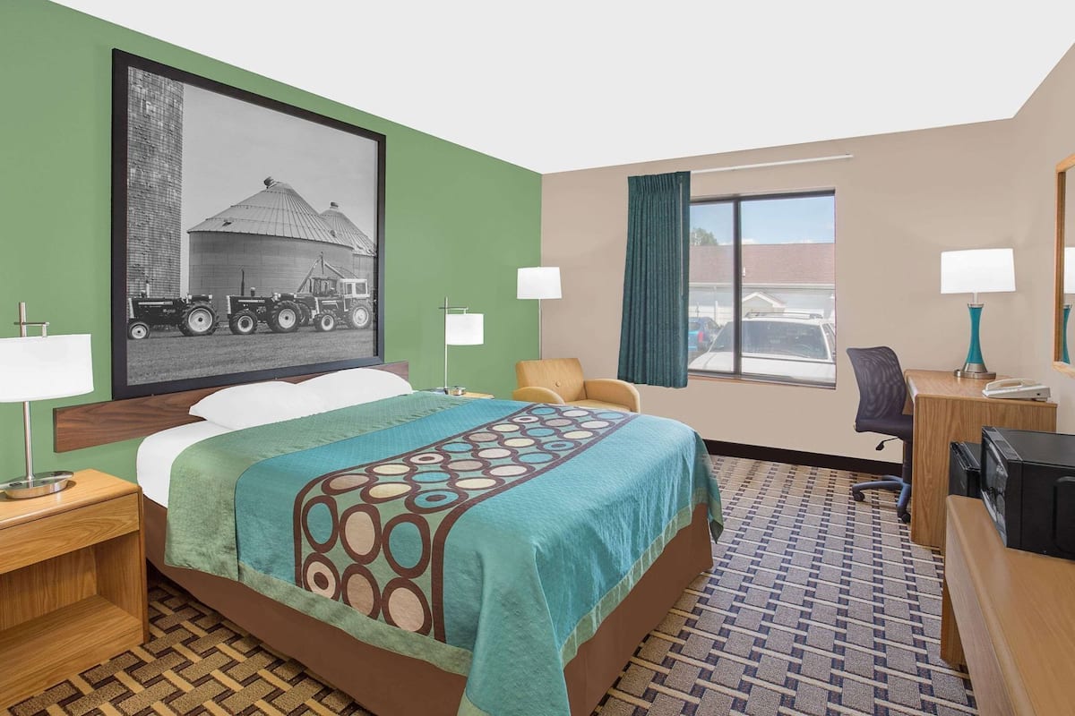 A hotel room with a bed, a desk, a chair, a desk lamp, a nightstand, a framed picture on the wall, and a window with curtains.