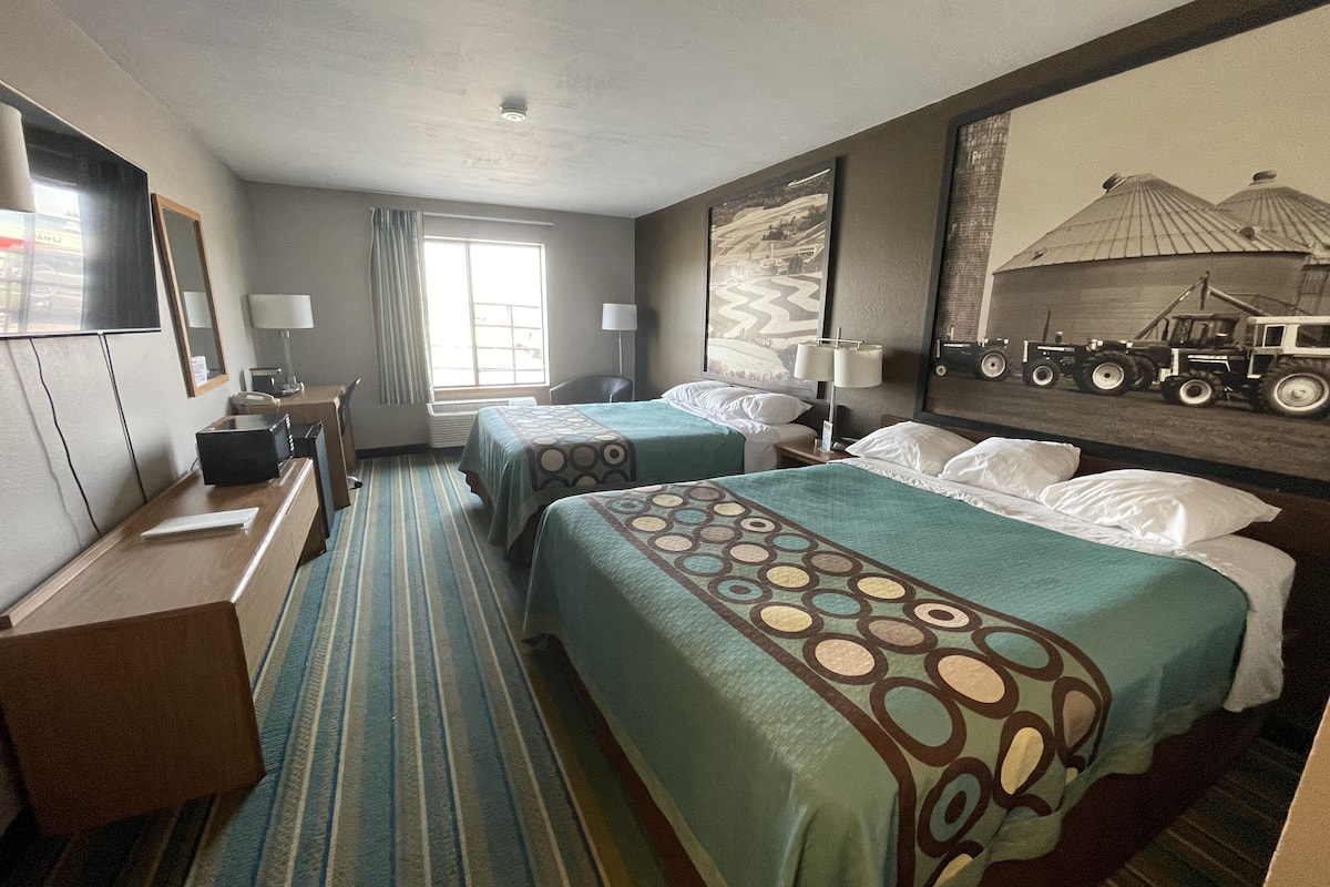 A hotel room with two beds, a desk, a chair, a TV, and a painting of tractors on the wall.