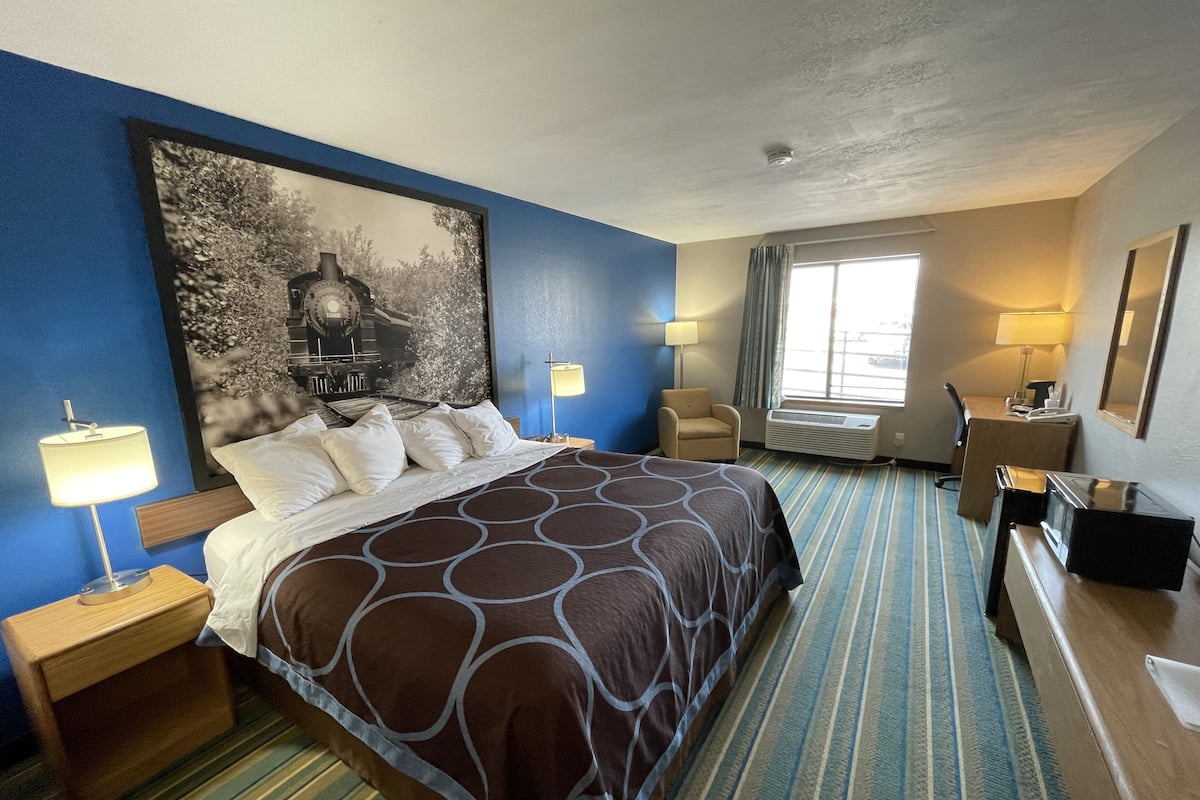 A hotel room with a large bed, a desk, a chair, a window, and a painting on the wall.