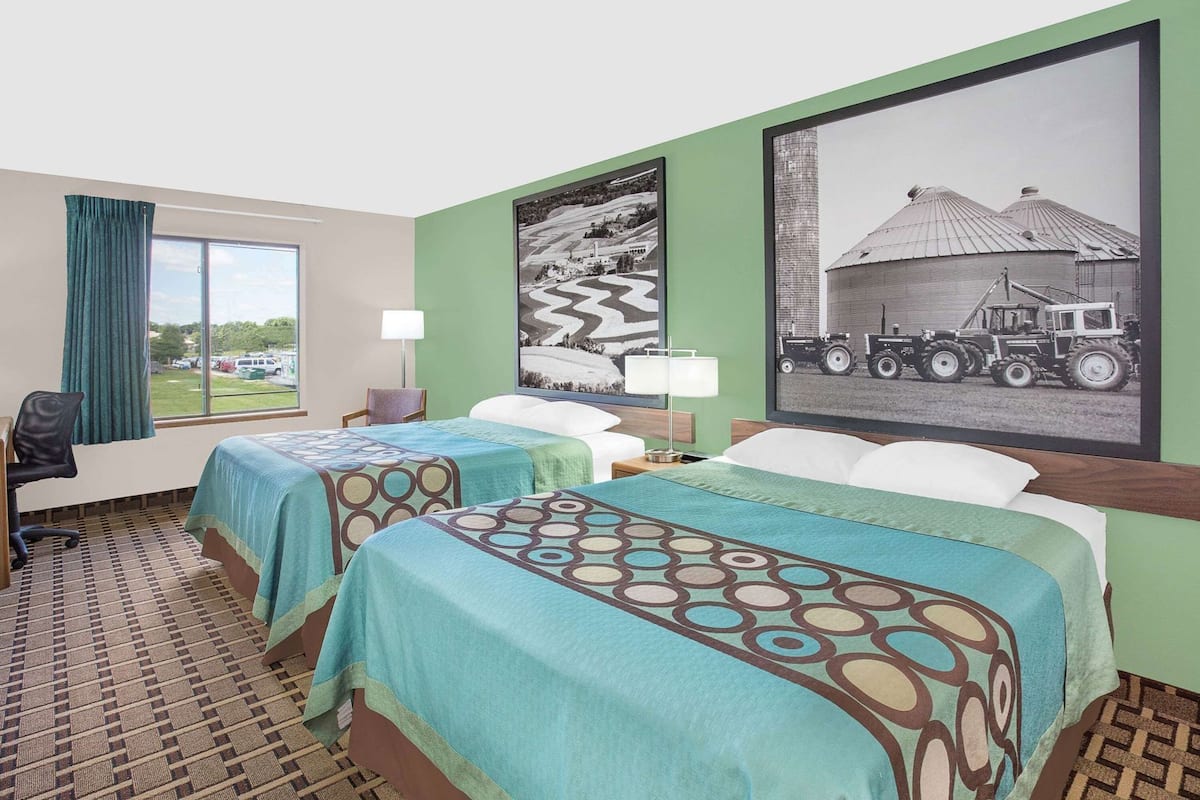 A hotel room with two beds, a desk, a chair, a window with curtains, and two wall-mounted lamps.